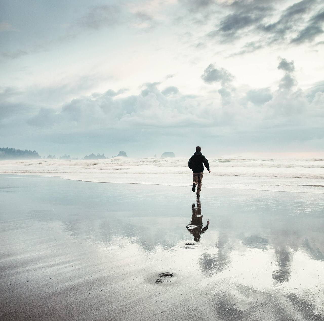A lone figure running across a wide reflective beach under an open sky