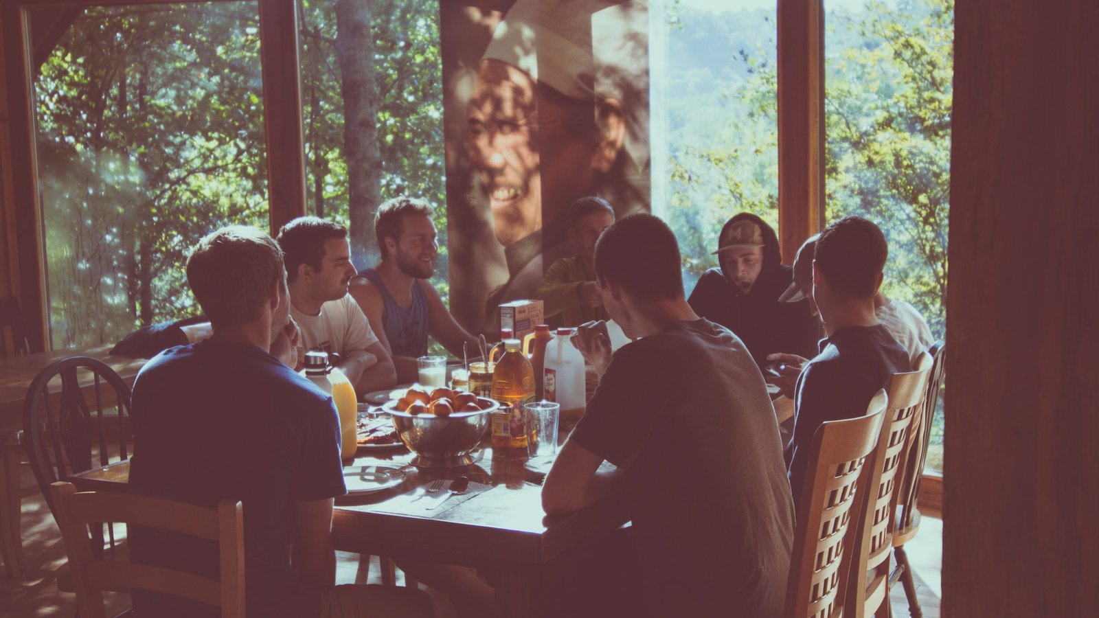 A group of friends sharing a long breakfast table in a room with tall windows looking out onto sunlit trees