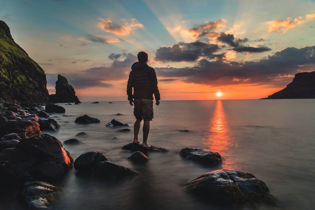 A lone figure standing on rocks looking out to sea at sunset