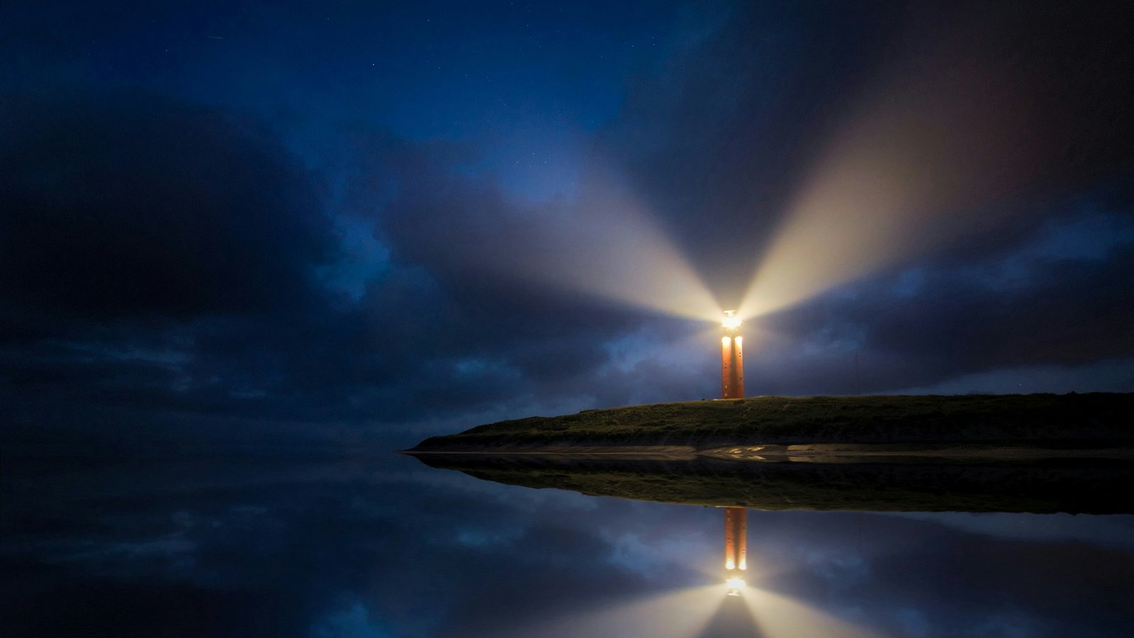 A lighthouse at night casting broad beams of warm light through cloud over a headland, mirrored in the still water below