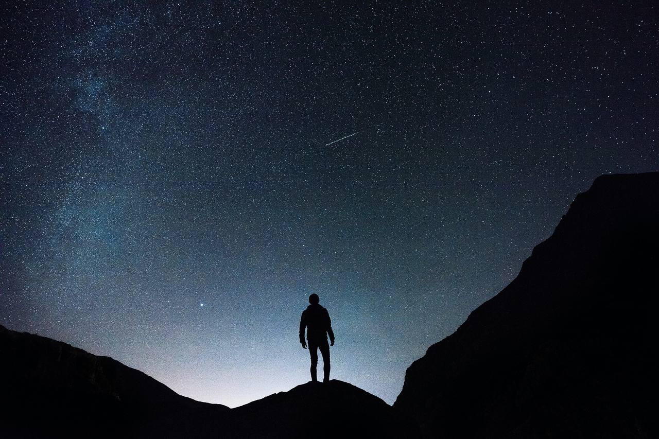 A lone figure standing on a ridge beneath the Milky Way and a vast field of stars